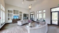 This single-floor space makes an immediate impression with its dramatic vaulted ceilings that lend an airy, expansive feel to the interior. A striking sculptural chandelier serves as a focal point overhead, adding a touch of refined elegance to the open layout. The main lounge area is furnished with plush, tufted sofas and a curved sectional arranged around a sleek metallic accent table, creating an inviting setting for conversation and relaxation. Light wood-toned flooring runs throughout, complementing the soft, neutral color palette of warm whites and greiges that defines the space. Built-in shelving niches are set into the walls, offering both decorative display space and a mounted flat-screen television. Floor-to-ceiling windows along one wall flood the interior with generous natural light while framing views of lush greenery outside. Glass-panel doors provide a seamless visual connection to an adjacent area, enhancing the sense of openness and flow. Black window and door trim provides clean contrast against the bright walls, adding a contemporary architectural detail. Overall, the space balances modern sophistication with comfortable, functional design, making it well-suited as a welcoming communal or gathering area.
