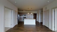 This single-floor residence showcases a contemporary open-concept layout centered around a well-appointed kitchen. The kitchen features a central island with a white base and light stone countertop, equipped with a sink and offering additional workspace or casual seating. Stainless steel appliances, including a refrigerator, range, and microwave, are complemented by medium-toned cabinetry that extends to the upper walls, providing generous storage. A granite or stone-style backsplash adds texture and visual interest behind the range.

Overhead, pendant lights hang above the island, creating focused task lighting, while a track lighting fixture illuminates the broader kitchen area. The ceilings are a generous height, contributing to an open and airy feel throughout the space.

Dark hardwood flooring flows continuously across the main living area, grounding the interior with warmth and contrast against the white walls and cabinetry. Multiple doors visible from the main space suggest access to additional rooms, including what appears to be a main entry door with a warm wood finish. The overall ambiance is clean, modern, and move-in ready.