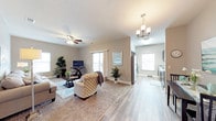 This single-floor space showcases an open-concept design that seamlessly connects the living area to the kitchen. The living room is finished with plush carpeting and features a ceiling fan with integrated lighting, creating a comfortable and functional atmosphere. Large windows allow natural light to fill the space, enhancing the warm, neutral color palette of the walls and trim. The living area comfortably accommodates seating arrangements centered around a coffee table, with space for a media setup along one wall. Sliding glass doors provide access to an outdoor balcony, extending the living space further. Transitioning into the adjoining area, the flooring shifts to smooth hardwood-style planks that carry through to the kitchen. The kitchen is well-appointed with stainless steel appliances, including a refrigerator, and features light-toned cabinetry with stone-look countertops. An elegant chandelier hangs overhead, adding a refined touch to the space. Throughout the unit, decorative greenery and thoughtful staging highlight the versatility and welcoming ambiance of this well-designed, single-level layout.