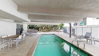 Located at 7513 Fountain Avenue in the heart of Los Angeles, this communal amenity space offers a well-appointed covered pool and spa area designed for relaxation and recreation. The centerpiece is a generously sized lap pool with clear turquoise water, complemented by a separate spa/hot tub, both finished with decorative tile trim and accessible via stainless steel handrails. The entire area is sheltered beneath a substantial concrete overhead structure supported by bold exposed beams, providing shade while remaining open to the surrounding greenery and natural light on multiple sides. The pool deck is finished in neutral stone-toned tile, offering a clean and durable surface throughout. A dedicated lounge area features modular seating and low tables, ideal for unwinding poolside. An outdoor dining setup with a wooden table and white chairs provides space for gatherings and meals. A gas BBQ grill station adds further functionality for entertaining. Lush, mature landscaping lines the perimeter fence, creating a private and serene atmosphere. The space is enclosed by a metal railing fence and accessed through a secured door, ensuring safety and controlled entry. Safety signage and pool rules are prominently displayed. This amenity space delivers a practical yet comfortable outdoor retreat in a prime Los Angeles location.
