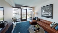This single-floor residence presents a thoughtfully designed open layout with a distinctly modern aesthetic. The living area serves as the centerpiece of the space, anchored by large floor-to-ceiling windows and a glass sliding door that frame expansive city and hillside views, flooding the room with natural light. A leather sectional sofa, round glass coffee table, and deep teal area rug contribute to a polished yet comfortable atmosphere.

The kitchen is well-equipped and efficiently laid out in a U-shape configuration, featuring light wood cabinetry with matte black hardware, white quartz countertops, a marble-style tile backsplash, and stainless steel appliances including a gas range and built-in microwave. Light wood flooring and recessed lighting enhance the clean, contemporary feel.

The primary bedroom offers generous proportions with floor-to-ceiling windows providing urban views and elegant draping curtains for privacy. A secondary bedroom maintains the same neutral palette with a window overlooking surrounding buildings and direct access to the hallway.

The bathroom is finished with large-format neutral floor tiles, a glass-enclosed walk-in shower with stone tile surround, a light wood vanity with white countertop, and a wide mirror, delivering a clean, spa-like environment consistent with the overall design language of the space.