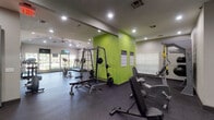 This single-level fitness center offers a comprehensive workout environment designed to support a wide range of exercise routines. The space is divided into distinct functional zones, each catering to different training needs. The cardio area is positioned near large windows that frame views of an outdoor pool and surrounding greenery, providing natural light and an open, airy atmosphere. Treadmills and an elliptical machine are positioned to take advantage of these views, accompanied by wall-mounted televisions for entertainment during workouts.

The strength training area features a prominent power rack with weight plates, a flat bench, and a cable machine, set against a bold lime-green accent wall that adds visual energy to the space. Motivational words and phrases are displayed across one wall, reinforcing an encouraging atmosphere. TRX suspension straps, stability balls, and a dedicated dumbbell rack with a graduated set of free weights further expand training options.

The flooring throughout consists of durable rubber gym matting, providing both safety and noise reduction. Recessed lighting illuminates the entire space evenly, while a ceiling fan supports ventilation. A French-style glass door connects the fitness center to an adjacent interior space, offering convenient access to the broader facility.