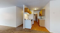 This single-floor residential unit offers a practical and well-organized layout across several distinct living areas. The kitchen is the functional heart of the home, equipped with warm honey-toned oak cabinetry both above and below the counters, a white electric range with overhead ventilation, a full-size refrigerator, and a double-basin sink. Wood-look flooring runs throughout the kitchen, adding warmth and durability to the space. A structural column partially divides the kitchen from the adjacent living area, which features plush wall-to-wall carpeting and a ceiling fan with integrated lighting. The open layout allows for flexible furniture arrangements. The bedrooms are generously sized, each finished with soft carpeting, baseboard heating units, and windows fitted with horizontal blinds that allow natural light to filter in. One bedroom offers a view of mature trees, while another overlooks a neighboring building exterior. The bathroom is neatly appointed with a single-sink vanity featuring wood cabinetry, a full tub and shower combination with a white surround, and wood-look flooring. A closet with sliding doors provides additional storage. Textured ceilings are present throughout, and the neutral color palette creates a clean, move-in ready environment.