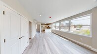 This thoughtfully designed single-floor unit offers a clean, contemporary aesthetic with an open-concept layout that seamlessly connects the living area to the kitchen. Light gray hardwood-style flooring runs throughout, complementing the neutral warm-beige walls and crisp white trim to create a cohesive, modern atmosphere.

The expansive living area is bathed in natural light, courtesy of a generous bank of large double-hung windows that frame views of the surrounding neighborhood streetscape. A wall-mounted mini-split unit provides efficient climate control, while recessed lighting ensures consistent illumination throughout the space.

The kitchen is a standout feature, showcasing white shaker-style cabinetry, striking granite countertops with bold veining, and a central island that doubles as a breakfast bar. Stainless steel appliances — including a range, microwave, and dishwasher — add a polished, functional touch. A dedicated window in the kitchen brings in additional natural light.

The bedroom is a calm, well-proportioned retreat with its own window offering neighborhood views and the same warm flooring found throughout the unit. The bathroom is clean and functional, fitted with a glass-enclosed walk-in shower, built-in shelving, and modern fixtures. Additional doors lead to utility and closet spaces, completing this well-appointed, move-in-ready unit.