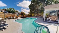 Located at 3214 Brassfield Rd in Greensboro, North Carolina, this outdoor amenity area centers around a generously sized freeform swimming pool with crystal-clear turquoise water. The pool features multiple entry points, including wide steps and stainless steel handrails, and is surrounded by an expansive concrete sundeck that offers ample room for lounging and socializing.

The deck is well-appointed with a variety of seating options, including traditional lounge chairs, woven hanging egg chairs, and umbrella-shaded seating areas, catering to both sunbathers and those seeking shade. A rustic wood-panel retaining wall runs along one side of the pool area, providing a natural, textured backdrop and adding warmth to the outdoor aesthetic. Mature trees frame the space, offering natural shade and a lush, green canopy overhead under open skies.

Adjacent to the pool is a single-story clubhouse building with a gabled roof, dark exterior siding, and white-trimmed French doors that open directly onto a covered porch with additional seating. The entire pool area is enclosed by a black iron security fence, ensuring safety and privacy. The combination of thoughtful landscaping, varied seating, and well-maintained facilities makes this an inviting and functional outdoor retreat.