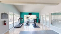 Located at 715 Marie Antoinette St. in Lafayette, Louisiana, this fitness center is a bright and functional exercise space designed to support a comprehensive workout routine. The room features vaulted ceilings with exposed beams and recessed lighting, creating an open and airy atmosphere that enhances the sense of space. A bold teal accent wall serves as a striking focal point, adding a vibrant energy to the otherwise clean, white interior.

Large sliding glass doors and generously sized windows with white blinds line the walls, flooding the space with natural light and offering direct views of an outdoor pool and patio area. Light-colored ceramic tile flooring covers the main area, complemented by dark rubber exercise mats that define the workout zones.

The cardio section is outfitted with a treadmill, elliptical trainer, and stationary and recumbent bikes. A wall-mounted flat-screen television above the teal accent wall provides entertainment during workouts. A dedicated strength training area features a multi-function cable machine and a weight bench, with full-length mirrors amplifying the sense of depth. A water cooler and additional signage are also present. A separate entry area with glass doors provides easy access to the outdoors, making this fitness center a well-rounded and inviting facility.