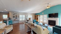 The Wyndamere clubhouse is a single-story gathering space designed to foster community and comfort. The open-concept layout is anchored by rich hardwood flooring that flows throughout the main living areas, complemented by neutral gray walls and bold teal accent walls that lend a coastal, energetic character to the space.

The main lounge area is expansive, featuring vaulted ceilings with ceiling fans and abundant natural light streaming through multiple white-framed windows with blinds. A striking fireplace with a white surround serves as a focal point, topped by a wall-mounted television and flanked by French doors that open to an outdoor deck or patio, offering views of the surrounding grounds. The lounge accommodates multiple seating arrangements, creating flexible spaces for socializing or relaxing.

Adjacent to the lounge is a bright dining nook with large windows and a glass-top table, as well as a fully equipped kitchen featuring wood cabinetry, black appliances, and tile flooring. A coffee station with bar seating adds a welcoming, communal touch. A secondary sitting area with tufted white furnishings and a chevron-front wood cabinet provides a quieter retreat within the space.