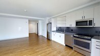 This studio residence offers a thoughtfully designed open-concept layout that seamlessly integrates the kitchen and living areas into a single, functional space. The kitchen is a standout feature, equipped with stainless steel appliances including a refrigerator, gas range, and built-in microwave. Crisp white cabinetry with open shelving provides ample storage, while dark countertops and a slate-blue subway tile backsplash add a contemporary contrast. A stainless steel sink with a modern faucet completes the well-appointed kitchen. Warm-toned hardwood flooring extends throughout the entire space, adding visual continuity and a sense of warmth. The open living area benefits from a clean, minimalist aesthetic with smooth white walls and ceilings, creating a bright and airy atmosphere. The entryway is clearly defined, featuring a solid entry door and additional interior door access. The overall design balances modern finishes with practical functionality, making efficient use of the open floor plan while maintaining a polished, contemporary look throughout.