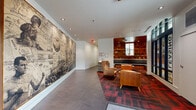 Located at 8338 North Interstate Avenue in Portland, Oregon, this single-floor lounge and hallway space serves as a dynamic communal gathering area for residents. The layout follows an angled, L-shaped configuration that seamlessly connects the building's entrance to its interior corridors.

The lounge area is anchored by a built-in L-shaped banquette upholstered in charcoal gray fabric, framed by warm wood paneling that extends up to a rich wood-clad ceiling. Recessed lighting illuminates the space, highlighting floor-to-ceiling built-in shelving filled with books and décor. A rustic reclaimed wood coffee table on casters serves as a natural centerpiece, grounding the seating nook with an earthy, textured element. Bold red-and-black plaid carpeting adds a strong visual identity throughout the seating zone.

The entry area features a large-format mural — a striking black-and-white collage celebrating Portland's history and culture — commanding an entire wall and immediately establishing a sense of place. Comfortable cognac leather chairs are arranged near glass-paneled storefront windows, allowing natural light to filter in from the street.

The hallway extends cleanly with dark tile flooring, crisp white walls, recessed lighting, and built-in mailboxes, offering a well-organized and functional passage. Together, these spaces reflect a thoughtfully designed, community-focused environment rooted in Pacific Northwest character.