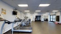 Located at 300 Tresser Blvd in Stamford, Connecticut, this fitness center offers a well-organized, single-floor layout designed to support a full range of workout routines. The space features a clean, functional aesthetic with light gray walls, a drop-panel ceiling equipped with bright fluorescent lighting, and durable rubber flooring throughout — ideal for high-traffic fitness use.

The cardio area is prominently featured along one side of the room, housing a row of treadmills and elliptical machines, including the T80 model treadmill. A wall-mounted flat-screen television is positioned for convenient viewing during workouts. The open floor plan allows for easy movement between equipment zones.

Toward the rear of the space, a dedicated free weights and strength training area is visible, complete with a dumbbell rack and additional equipment. Mirrored wall panels in this section enhance the sense of space and allow users to monitor their form during exercises. A doorway leads to an adjoining area, suggesting additional utility or storage space.

The fitness center is well-lit, spacious, and thoughtfully arranged to accommodate both cardiovascular and strength training needs, making it a comprehensive facility for everyday fitness use.