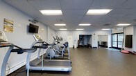 Located at 300 Tresser Blvd in Stamford, Connecticut, this fitness center offers a well-organized, single-floor layout designed to support a full range of workout routines. The space features a clean, functional aesthetic with light gray walls, a drop-panel ceiling equipped with bright fluorescent lighting, and durable rubber flooring throughout — ideal for high-traffic fitness use.

The cardio area is prominently featured along one side of the room, housing a row of treadmills and elliptical machines, including the T80 model treadmill. A wall-mounted flat-screen television is positioned for convenient viewing during workouts. The open floor plan allows for easy movement between equipment zones.

Toward the rear of the space, a dedicated free weights and strength training area is visible, complete with a dumbbell rack and additional equipment. Mirrored wall panels in this section enhance the sense of space and allow users to monitor their form during exercises. A doorway leads to an adjoining area, suggesting additional utility or storage space.

The fitness center is well-lit, spacious, and thoughtfully arranged to accommodate both cardiovascular and strength training needs, making it a comprehensive facility for everyday fitness use.