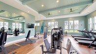 This fitness center offers a generously sized, single-floor layout designed to accommodate a full range of workout activities. The space is defined by its coastal-inspired aesthetic, featuring soft seafoam green walls, light hardwood-style flooring, and a distinctive coffered ceiling finished in a complementary teal tongue-and-groove wood detail. Dual ceiling fans and recessed lighting throughout ensure comfort and visibility across the entire facility.

The main workout area is well-appointed with a comprehensive selection of strength training machines, including cable and multi-station resistance units, adjustable benches, free weights, and a dumbbell rack. Cardio equipment such as treadmills and elliptical machines lines the perimeter, with large windows providing ample natural light and outdoor views. Full-length mirrors along one wall visually expand the space and support form-conscious training.

A raised studio alcove houses a row of spin bikes, stability balls in multiple sizes, and storage for stand-up paddleboards — a distinctive feature that reflects an active, recreation-oriented lifestyle. A wall-mounted flat-screen TV and camera tripod suggest the space supports virtual and on-demand fitness programming. The overall ambiance is bright, open, and energizing, catering to diverse fitness needs.