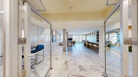 Located at 461 Railroad Ave in Westbury, NY, this elegantly designed amenity lounge at The Cornerstone Westbury is a generously sized, single-floor social hub that seamlessly blends luxury aesthetics with versatile functionality. The space is unified throughout by striking marble-look tile flooring, marble-clad columns, and crisp white wainscoting that lend a refined, upscale character to every zone.

At the heart of the lounge is a stunning bar area, where a richly veined stone countertop is paired with gold-accented bar stools and illuminated by a row of sleek pendant lights. Open shelving behind the bar displays decorative accessories alongside a coffee station. Adjacent to the bar, a high-top dining table bathed in natural light from large windows is crowned by a distinctive teal glass chandelier, creating an inviting spot for casual gatherings.

The lounge area features a soaring marble fireplace surround anchoring a flat-screen TV, flanked by plush seating and large windows that draw in abundant natural light and outdoor views. Curated large-format artwork adorns the walls throughout, adding a gallery-like ambiance. A shuffleboard table provides a recreational focal point, while glass-walled private meeting rooms with contemporary chandeliers and quilted lounge chairs offer quieter, flexible spaces for focused gatherings or collaborative work.