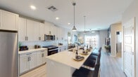 This well-designed single-floor residence offers a thoughtfully planned open-concept layout that seamlessly connects the kitchen, dining, and living areas. The kitchen is a standout feature, showcasing white shaker-style cabinetry extending to the ceiling, a herringbone tile backsplash, quartz countertops, stainless steel appliances including a gas range, microwave, refrigerator, and dishwasher, and a generously sized center island with bar seating illuminated by pendant lighting. Light wood-style flooring runs throughout the main living areas, adding warmth and continuity. The living room is bright and airy, benefiting from natural light streaming through large sliding glass doors that open to a private balcony with open views. The bedrooms feature plush carpeting, ceiling fans, and large windows that invite natural light. The primary suite connects to a well-appointed bathroom featuring a double vanity with backlit mirrors, quartz countertops, white cabinetry with matte black hardware, and direct access to a dedicated walk-in closet. A separate in-unit laundry area with side-by-side washer and dryer adds everyday convenience. The overall aesthetic is clean, contemporary, and cohesive throughout.