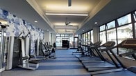 This fitness center offers a well-appointed workout environment designed with both functionality and aesthetics in mind. The elongated layout provides ample space for a diverse range of equipment, including a row of treadmills along one side and a selection of strength-training machines along the other, allowing users to move freely between stations without congestion.

The interior design is a standout feature, highlighted by a bold blue-and-white abstract accent wall that adds a dynamic visual element to the space. Striped blue and grey carpeting complements the color palette, reinforcing the modern, clean aesthetic throughout.

Lighting is a key component of the atmosphere, with recessed ceiling fixtures and long linear LED lights providing bright, even illumination across the room. Ceiling fans are evenly spaced along the length of the ceiling, contributing to air circulation and comfort during workouts.

Large windows along one side of the room flood the space with natural light and offer views to the outdoors, creating an open and energizing environment. At the far end, glass doors lead to an additional area, enhancing the sense of depth and accessibility within the facility.