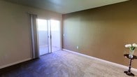 This single-floor space presents a straightforward and functional layout. The primary room visible is finished with wall-to-wall carpet in a neutral beige-gray tone, providing a soft underfoot feel throughout the space. The walls are painted in a warm tan hue with a lightly textured finish, complemented by clean white baseboards that frame the perimeter of the room.

A prominent sliding glass door serves as the focal point of the room, fitted with sheer white curtains that allow abundant natural light to fill the interior. The sliding door provides direct access to an outdoor balcony area, which features a tiled floor and a railing, offering a seamless connection between the indoor and outdoor areas.

The room is open and uncluttered, with a practical layout that allows for flexible furniture arrangement. Electrical outlets are positioned along the walls, ensuring utility throughout the space. The overall ambiance is clean, bright, and understated, with a neutral color palette that provides a versatile backdrop suitable for a variety of interior styles.