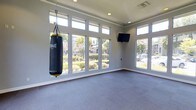 This dedicated yoga and fitness room is a bright, well-appointed space designed to support a variety of exercise and wellness activities. The room is flooded with natural light through a generous arrangement of floor-to-ceiling windows and transom windows above, offering views of lush greenery and outdoor landscaping beyond. The expansive glazing creates an open, airy atmosphere that enhances the sense of space throughout.

The floors are covered in durable, cushioned interlocking fitness tiles in a neutral gray tone, providing a comfortable and supportive surface suited for yoga, stretching, bodyweight training, or high-impact workouts. A heavy-duty Everlast punching bag is suspended from a ceiling-mounted chain, adding a functional element for cardio and strength conditioning.

A wall-mounted flat-screen television is positioned at an ideal viewing height, allowing users to follow along with workout videos or fitness classes during their sessions. The walls are finished in a clean, soft gray with white crown molding and baseboards, contributing to a calm and focused environment. Recessed lighting and a ceiling vent ensure year-round comfort. The overall design of the room balances practicality with a welcoming ambiance, making it a versatile space for both energetic training and mindful practice.