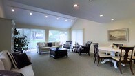 This single-floor space offers a welcoming and well-appointed interior defined by its striking vaulted ceilings, which lend a sense of openness and volume to the main living area. The space is bathed in natural light, thanks to a generous arrangement of large windows and vertical blinds that span nearly the full length of the exterior wall, offering pleasant views of the surrounding greenery outside.

The living area is thoughtfully designed, featuring a neutral-toned carpet that complements the light-colored walls and creates a warm, comfortable atmosphere. A combination of track lighting and recessed ceiling fixtures ensures excellent illumination throughout the day and evening. The living space flows naturally into a dining area, making the layout ideal for both relaxed everyday living and entertaining guests.

Furnishings in the space reflect a classic, coordinated aesthetic — a plush sofa and decorative accent chairs with patterned cushions are arranged around a dark wood coffee table, while a round dining table and matching chairs anchor the adjacent dining zone. The interplay of dark wood tones against the light carpet and walls creates an elegant visual contrast. Overall, the space presents a refined yet inviting ambiance suited to a variety of lifestyles.