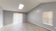 This single-story space is defined by its vaulted ceilings, which lend an airy, open quality to the main living area. A centrally positioned skylight floods the room with natural light, complementing the light wood-style flooring that runs throughout. The neutral gray walls provide a clean, contemporary backdrop, while vertical blinds on a large sliding door opening further enhance brightness and offer access to the outdoors.

The living area is generously proportioned, with enough room to accommodate a variety of furniture arrangements. A pass-through opening connects it seamlessly to the kitchen, which makes a bold visual statement with its dark espresso cabinetry extending from floor to ceiling. The kitchen is equipped with black appliances, including a range, dishwasher, and refrigerator, alongside a double-basin sink and ample counter space with a speckled laminate finish.

The bathroom is neatly appointed, featuring a full tub-shower combination with white tile surround, a toilet, and a vanity with a speckled granite-style countertop, dark cabinetry, and a large mirror above. Additional rooms, visible in the floorplan, include a carpeted bedroom, offering a comfortable contrast to the hard-surface flooring elsewhere. Overall, the space presents a cohesive, modern design with practical finishes throughout.
