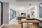 Located at 32 SE Second Ave in the heart of Delray Beach, Florida, this beautifully appointed single-floor amenity space offers a sophisticated and functional environment designed for community and collaboration. Light wood-look flooring flows seamlessly throughout, unifying the various zones and contributing to the space's warm, modern aesthetic.

The welcoming reception area features a striking white island counter adorned with a vibrant living moss display, flanked by wire chairs and crowned by an elegant tiered glass chandelier. Built-in shelving on either side provides both storage and visual depth, while a cozy built-in bench with integrated bookshelves adds a comfortable reading nook.

A central conference and lounge zone is defined by a sleek white communal table illuminated by a cluster of Edison pendant lights and divided from the lounge by a sculptural white vertical slat partition. The lounge area features ample seating, a wall-mounted TV, and floor-to-ceiling glass sliding doors that open to an outdoor terrace, flooding the space with natural light.

The kitchenette is outfitted with gray shaker cabinetry, stainless steel appliances, a matte black faucet, and white subway tile backsplash. A hallway with soft blue-gray walls leads to a stainless steel elevator, completing this well-rounded and thoughtfully curated space.