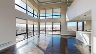 This impressive two-floor loft-style space showcases a bold industrial-modern design that balances open living with thoughtful details. The main living area commands attention with its dramatic double-height ceilings, exposed silver ductwork, and an expansive wall of floor-to-ceiling windows framed in dark steel. These oversized windows span both levels, flooding the space with abundant natural light and offering sweeping panoramic views of the surrounding cityscape and distant landscape. Rich, dark hardwood flooring runs throughout the main living areas, reflecting sunlight and adding warmth to the open-plan layout. A staircase connects the two floors, with the upper level featuring a mezzanine-style overview of the soaring main space below. The kitchen is sleek and contemporary, appointed with marble-look quartz countertops, a bold geometric dark tile backsplash, flat-front cabinetry in a two-tone finish, and a full suite of stainless steel appliances. The bathroom is clean and minimalist, finished with large-format neutral floor tiles, white subway tile in the walk-in shower, and matte black hardware accents. Bedroom areas are carpeted and bathed in natural light, offering a calm contrast to the dramatic main living space. Throughout, the design strikes a refined balance between industrial character and modern comfort.