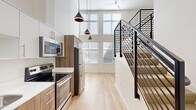 Located at 1670 SW Alder St in Portland's vibrant West End neighborhood, this modern multi-level unit showcases an efficient and thoughtfully designed layout. The main level opens to a contemporary kitchen equipped with stainless steel appliances, including a French door refrigerator, electric range, and built-in microwave. Wood-toned cabinetry paired with crisp white countertops and light hardwood flooring creates a clean, modern aesthetic throughout. A defining architectural feature is the dramatic double-height ceiling in the main living area, which amplifies the sense of space and is accentuated by an impressive wall of large, multi-pane windows that flood the interior with abundant natural light. Sleek cone-shaped pendant lights descend from above, adding a stylish focal point. A bold black steel staircase with horizontal rail balusters leads to the upper level, where carpeted flooring, recessed lighting, and a ceiling fan contribute to a comfortable, quieter retreat. The upper level also includes a closet and open landing area. A dedicated laundry nook houses a stacked washer and dryer unit, adding everyday convenience. The bathroom completes the functional offerings of this compact yet striking space.