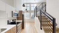 Located at 1670 SW Alder St in Portland's vibrant West End neighborhood, this modern multi-level unit showcases an efficient and thoughtfully designed layout. The main level opens to a contemporary kitchen equipped with stainless steel appliances, including a French door refrigerator, electric range, and built-in microwave. Wood-toned cabinetry paired with crisp white countertops and light hardwood flooring creates a clean, modern aesthetic throughout. A defining architectural feature is the dramatic double-height ceiling in the main living area, which amplifies the sense of space and is accentuated by an impressive wall of large, multi-pane windows that flood the interior with abundant natural light. Sleek cone-shaped pendant lights descend from above, adding a stylish focal point. A bold black steel staircase with horizontal rail balusters leads to the upper level, where carpeted flooring, recessed lighting, and a ceiling fan contribute to a comfortable, quieter retreat. The upper level also includes a closet and open landing area. A dedicated laundry nook houses a stacked washer and dryer unit, adding everyday convenience. The bathroom completes the functional offerings of this compact yet striking space.
