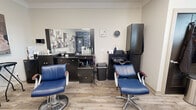 This thoughtfully designed salon space offers a complete, multi-service environment within a compact yet efficiently organized single-floor layout. The main styling area features two professional hydraulic chairs upholstered in rich blue leather, positioned in front of a dark espresso vanity with an integrated mirror, ample storage drawers, and a full array of styling tools and products. Adjacent to the styling stations, a dedicated shampoo station includes a built-in bowl and a reclined chair, providing a seamless workflow for hair care services.

A separate zone within the space serves dual purposes, housing a hooded dryer chair and a nail or esthetics workstation. This area is distinguished by a bold blue damask accent wall that adds a striking design contrast to the otherwise neutral, warm-toned palette. Two windows with white blinds fill the space with natural light, and small potted plants on the sills lend a welcoming, organic touch.

Throughout, light wood-look flooring anchors the interior with a clean, contemporary feel, while crown molding along the ceiling adds a refined architectural detail. Curated artwork, including a prominent abstract triptych in blush and muted tones, decorates the walls, enhancing the salon's polished and inviting ambiance. A glass display shelf near the entry showcases retail products, completing the professional presentation of this well-equipped salon.