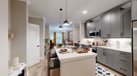 Located at 5672 Peachtree Pkwy in Peachtree Corners, GA, this thoughtfully designed one-bedroom, one-bathroom residence offers a cohesive open-concept layout that maximizes its well-proportioned footprint. The kitchen is a standout feature, showcasing gray shaker-style cabinetry, stainless steel appliances including a gas range and side-by-side refrigerator, white quartz countertops, and a central island with seating — all illuminated by stylish pendant lighting. The kitchen flows seamlessly into the living room, which is distinguished by crown molding, a warm terracotta accent wall, a modern chandelier, and large windows dressed in gold curtains that invite natural light. Light-toned hardwood-style flooring runs throughout the main living areas, adding warmth and continuity. The bedroom offers a cozy retreat with plush carpeting, a blue accent wall, a natural light window, and a ceiling fan. The bathroom features gray cabinetry, a white quartz vanity, black hardware accents, and in-unit laundry conveniently situated nearby. A dedicated closet provides additional storage. Crown molding, recessed lighting, and a refined neutral palette tie the entire space together with a polished, modern aesthetic.