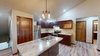 This single-floor residence offers a cohesive and functional layout designed with modern finishes and warm, natural tones throughout. The kitchen serves as a standout centerpiece, featuring rich dark wood cabinetry, light granite countertops, and a full suite of stainless steel appliances including a refrigerator and electric range. A peninsula with a double basin sink and an eye-catching industrial-style pendant light fixture adds both style and practicality to the space. Recessed lighting further brightens the kitchen area, which benefits from vaulted ceiling lines that enhance the sense of openness.

The living and dining areas flow naturally from the kitchen, unified by durable laminate wood-look flooring and neutral wall tones that provide a versatile backdrop. A decorative chandelier anchors the dining space, while natural light filters in through windows that offer views of the surrounding greenery.

The bedrooms are well-sized, with one featuring a ceiling fan, window with outdoor views, and a wall-mounted AC unit. Warm-toned wood double doors provide closet access in another bedroom. The bathroom is clean and functional, with a dark wood vanity, granite countertop, and a shower-tub combination. Additional storage is available through a dedicated walk-in closet space with built-in shelving. Warm wood interior doors and rich baseboards add a consistent, quality touch throughout.