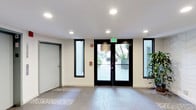 This single-floor space presents a clean and functional lobby area designed with modern commercial sensibilities. The entry is anchored by a set of black-framed double glass doors, flanked by tall, narrow sidelights that invite an abundance of natural light into the interior. Additional narrow windows on either side of the entryway further brighten the space, offering glimpses of exterior landscaping that includes greenery and a paved walkway outside.

The ceiling is finished in white and outfitted with a series of recessed can lights, providing even, well-distributed artificial illumination throughout. Safety features are clearly integrated into the design, including a prominently displayed green illuminated exit sign above the entry doors and a red fire alarm pull station mounted to the wall.

The flooring consists of large-format tiles in a neutral tone, with a decorative patterned tile inset near the entrance that adds a subtle design accent. The walls are finished in a light, neutral hue that contributes to the bright and open ambiance of the space. A secondary door with a gray frame is visible to one side, suggesting additional access points or utility spaces. The overall aesthetic is understated and practical, consistent with a professional or residential building lobby.