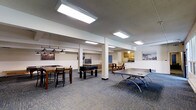 This single-floor recreational common space is thoughtfully designed to offer a variety of leisure and social amenities in one open, accessible layout. The main area is anchored by a full-size ping-pong table and a foosball table, providing ample entertainment options for residents or guests. A bar-height table with seating is positioned beneath stylish pendant lights, creating a casual dining or social nook. A comfortable lounge area with a sofa offers a relaxed spot to unwind between activities. The space is finished with durable, low-pile carpet in a neutral tone that complements the clean, white walls adorned with framed artwork, lending a polished yet welcoming feel. Structural columns divide the open floor plan subtly without interrupting the flow of the space. Bright, recessed fluorescent ceiling fixtures ensure the entire area is well-lit throughout. Toward the rear of the space, a compact kitchenette with cabinetry and countertops provides functional convenience for gatherings. A blue accent door adds a pop of color and leads to an adjacent area. Overall, the space balances functionality and comfort, making it an ideal communal hub for recreation and socializing.