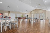 This versatile multi-use room in Montgomery, Alabama, offers a generously sized, single-level space designed to accommodate a wide range of activities and social gatherings. The open floor plan is anchored by warm hardwood-style flooring that flows seamlessly throughout, complemented by neutral, cream-toned walls and a combination of vaulted and textured ceilings fitted with recessed lighting.

The recreation area is a standout feature, providing ample room for a full-size pool table and a ping-pong table, making it well-suited for leisure and entertainment. Dining tables with cross-back chairs offer space for communal meals or group activities. A dedicated lounge area invites relaxation with comfortable armchairs arranged around glass-topped side tables, centered on a classic brick fireplace with a decorative mantel — a warm focal point for the room.

A small fitness corner features exercise equipment and benefits from natural light streaming in through multiple windows. Additional areas include a workspace with seating, a snack vending machine, and ample closet storage via built-in bifold doors. The space also connects to a secondary room with pendant lighting and additional seating arrangements. Overall, the layout and amenities make this an adaptable, welcoming environment suited for community engagement and recreation.