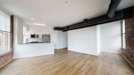 This single-floor loft-style unit masterfully combines industrial heritage with modern design. Throughout the space, warm light hardwood flooring runs continuously, complementing the striking contrast between crisp white walls and original exposed brick. Prominent black steel ductwork and structural beams trace the ceilings, reinforcing the building's industrial character while adding an architectural focal point.

The open-concept living area is generously proportioned and bathed in natural light through multiple large windows set within the exposed brick facade, offering views of the surrounding neighborhood. The kitchen is sleek and contemporary, featuring white cabinetry, a large white quartz island with an integrated sink, subway tile backsplash, and a full suite of stainless steel appliances including a refrigerator, microwave, and range.

The bedrooms continue the loft aesthetic, each showcasing exposed brick accent walls and tall windows that flood the rooms with natural light. Closet space is well integrated into each bedroom. The bathroom is finished to a modern standard, with large-format neutral tiles, a floating white vanity, and a glass-enclosed walk-in shower. The overall ambiance is airy, bright, and distinctly urban, with a thoughtful balance of raw industrial elements and refined contemporary details.