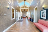 Located at 3688 Parkmoor Village Dr in Colorado Springs, CO, this impressive single-floor community clubhouse offers a generous and well-appointed gathering space designed for both comfort and functionality. The entryway makes an immediate statement with soaring vaulted ceilings, a dramatic clerestory that floods the corridor with natural light, textured stone-finished walls, and rich wood-look tile flooring that flows throughout the space. A central chandelier and wall sconces add warm, layered lighting to the grand entrance hall, which is lined with seating areas and framed views into adjacent rooms. The expansive main lounge is the architectural centerpiece, featuring striking exposed wooden ceiling beams, dual pendant chandeliers, and a floor-to-ceiling stone fireplace that anchors the room with rustic elegance. Large windows on multiple walls invite abundant natural light and offer views of the surrounding Colorado landscape. A bright, windowed sitting alcove provides an intimate retreat with vaulted ceilings and curated seating. The kitchen and dining area transition seamlessly from the main hall, offering pendant lighting, dining tables, and a service counter. Thoughtful art installations and carefully selected furnishings complete the welcoming, refined ambiance of this versatile community space.