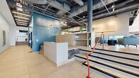 Located at 221 Monticello Ave in Williamsburg, Virginia, this expansive two-story facility offers a thoughtfully designed blend of fitness, social, and collaborative spaces totaling over 15,000 square feet across two floors. The welcoming ground-floor lobby greets visitors with large floor-to-ceiling glass entry doors, light wood flooring, a bold green ceiling accent, and a reception desk flanked by comfortable seating areas that bathe in natural light. A striking open-tread staircase with a dark steel structure connects the floors, adding an industrial-modern design element. The upper floor opens to a dramatic double-height atrium with an eye-catching wood-panel ceiling feature and integrated lighting, overlooking a lounge and bar area below. Flexible co-working spaces on the upper level feature slatted ceilings, expansive windows with outdoor views, collaborative table seating, and glass-walled meeting rooms. A dedicated conference room with wide-plank hardwood floors and panoramic windows seats a large group around a substantial central table. A game room with an exposed dark ceiling, industrial pendant lighting, a pool table, and lounge seating provides a relaxed social setting. The well-equipped exercise room features tile flooring, mirrored walls, and a comprehensive array of strength and cardio equipment. Additional rooms include a media lounge with birch cabinetry, a mounted display, and an exposed concrete accent wall, completing this versatile, amenity-rich facility.