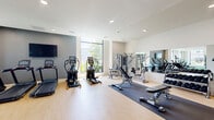 Located at 39 Trotter Road in Weymouth, MA, this fitness center is a comprehensive, single-floor facility designed to support a wide range of workout routines. The space is thoughtfully divided into distinct training zones, each catering to different fitness disciplines.

The main cardio area features a lineup of treadmills, elliptical trainers, and stationary bikes arranged along large floor-to-ceiling windows that flood the room with natural light and offer views of the surrounding greenery. Wall-mounted flat-screen televisions enhance the cardio experience. Light hardwood-style flooring throughout this zone gives the space a clean, modern aesthetic.

Adjacent to the cardio section is a dedicated strength training area outfitted with multi-station cable machines, adjustable benches, a full rack of dumbbells, and weight training equipment. Full-length mirrors line the walls, amplifying the sense of openness and allowing users to monitor their form.

A separate functional training room features green turf flooring and is equipped with a heavy punching bag, resistance training systems with TRX-style cables, spin bikes, rowing machines, and a variety of medicine balls and kettlebells — ideal for high-intensity and cross-training workouts.

Recessed lighting throughout, neutral wall tones, and glass partition elements contribute to the facility's bright, contemporary ambiance. The overall layout supports a seamless and versatile fitness experience.