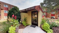 This single-floor rental office serves as the leasing hub for a residential community, offering prospective and current residents a convenient, accessible point of contact. The exterior of the building is clad in yellow brick tile and features a distinctive dark red awning prominently displaying the office name and contact number, giving it a recognizable and professional appearance. A concrete walkway leads directly to the dark-framed glass entry door, providing a clean and straightforward approach to the entrance.

The landscaping surrounding the office is notably well-maintained, with lush plantings of hostas, coneflowers, cannas, and other flowering plants arranged in mulched beds on either side of the entry path. This greenery adds color and warmth to the approach, creating an inviting atmosphere for visitors. Mature trees are visible in the background, contributing to a shaded, park-like setting within the broader community grounds.

The surrounding residential buildings, visible in the background, feature red brick construction with balconies, indicating a multi-story community layout. The rental office itself is a compact, standalone structure designed specifically to support leasing operations, offering a focused and efficient space for staff to assist residents and prospective tenants. Overall, the office presents a tidy, approachable, and well-kept environment reflective of the community it serves.