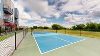 This outdoor recreational space features two dedicated pickleball courts, finished in a striking combination of bright blue and sage green playing surfaces with crisp white court markings. Each court is equipped with a regulation net, providing a complete and ready-to-play environment for residents and guests. The courts are fully enclosed by sturdy chain-link fencing, ensuring safety and containment during play while maintaining open sightlines to the surrounding landscape. Lush green grass borders the courts on multiple sides, and mature trees line the perimeter, offering a natural backdrop against wide-open skies. The facility is situated adjacent to a modern multi-story residential building, characterized by a contemporary mix of brick and horizontal siding with private balconies overlooking the courts. Additional community amenities, including what appears to be a pool area, are visible nearby, reinforcing the courts as part of a broader outdoor amenity offering. The open-air setting allows for abundant natural light throughout the day, and the expansive sky above creates an airy, energetic atmosphere well-suited for both casual play and competitive matches.