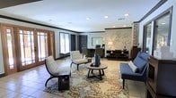 This single-floor space offers a thoughtfully designed interior that combines comfort and sophistication. The main lounge area serves as the centerpiece, featuring light-toned tile flooring accented by a richly patterned area rug in warm earth tones. A curated seating arrangement — including plaid accent chairs, a sleek navy sofa, and a round wooden coffee table — creates an inviting and functional gathering space. Recessed lighting illuminates the room from above, complemented by a decorative table lamp that adds warmth to the atmosphere. Crown molding and dark trim work frame the space with a refined architectural detail, while an exposed brick column introduces a touch of rustic character. The walls are decorated with an artful arrangement of circular wall plates and a floral accent piece, adding visual interest and texture. A wooden credenza with open shelving provides both display and storage space. Large doors with glass panels allow natural light to filter in, enhancing the open, airy feel of the interior. A dedicated desk area toward the back of the room suggests a workspace or reception function, making this space as practical as it is visually appealing.