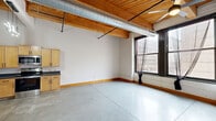 Located at 1110 Washington Ave in downtown St. Louis, this single-floor amenity space offers a well-rounded combination of social and fitness facilities. The layout is anchored by an open lounge area furnished with tan leather sofas arranged over a colorful area rug, complemented by a wooden table and chairs. A freestanding open shelving unit with metal framing and warm wood shelves adds a modern decorative element, while potted greenery introduces a touch of life to the space. Adjacent to the lounge, a wall-mounted mailbox bank and glass-paneled doors provide functional building amenity access.

The fitness center occupies a dedicated area with noticeably elevated ceilings, exposed ductwork, and track lighting. It is equipped with a treadmill, stationary spin bike, a full dumbbell rack, a TRX suspension training station, a weight bench, and a smart interactive mirror for guided workouts. Full-length mirrors line the fitness area walls, enhancing the sense of openness. Large interior windows connect the gym visually to the lounge area, maintaining a sense of connectivity throughout the floor. Neutral-toned carpet tiles, white walls, and industrial-style exposed mechanical systems give the space a clean, modern aesthetic consistent with an urban residential building's amenity offering.
