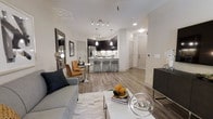 This single-floor residence showcases a thoughtfully designed open-concept layout that seamlessly connects the living, dining, and kitchen areas into one cohesive space. Light wood-grain flooring extends throughout, lending warmth and visual continuity to the interior.

The kitchen is a standout feature, equipped with dark cabinetry that contrasts sharply against light-toned walls and countertops. Stainless steel appliances, including a refrigerator, add a polished, modern touch. An island or peninsula with sleek bar-height stools provides casual seating and serves as a natural dividing element between the kitchen and the open living area. Pendant lighting hangs above this space, complementing the recessed and track lighting that illuminates the entire unit.

Adjacent to the kitchen, a compact bistro-style dining nook with a round table and accent chairs offers an intimate setting for meals. The living area features a contemporary grey sofa, decorative accessories, and a light-toned area rug that anchors the space. Neutral grey-white walls throughout create a clean, airy ambiance. The overall aesthetic is modern and cohesive, balancing functionality with refined, contemporary style.