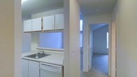 This single-floor residential space offers a clean, functional layout with a straightforward design. The kitchen is outfitted with white upper cabinetry, white countertops, a stainless steel sink, and a built-in dishwasher, presenting a practical and tidy cooking area. A partial wall divides the kitchen from the adjoining living space, maintaining an open feel while providing subtle definition between the two areas. The living area features wall-to-wall carpeting in a neutral tone, contributing to a comfortable, understated ambiance. A doorway visible from the kitchen leads further into the unit, suggesting a connected, flowing floor plan. Natural light enters through a tall, narrow window, brightening the space with a soft, cool glow that complements the light blue and white color palette found throughout. The walls are finished in a smooth, light texture, and the overall aesthetic is simple and neutral, lending itself well to personalization. The unit's single-floor layout ensures easy navigation, making the space feel cohesive and accessible throughout.
