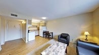 This single-floor unit is designed around an open-concept layout that seamlessly connects the living area and kitchen into one functional space. The flooring throughout is light wood laminate, lending a clean and cohesive look to the interior, while neutral beige walls create a bright, understated atmosphere.

The kitchen is positioned toward the rear of the space and is equipped with light-toned cabinetry, a white refrigerator, a stovetop range, and a sink with under-cabinet ventilation. Overhead fluorescent lighting ensures the kitchen area is well-lit for everyday use. A small dining set with a dark wood table and two chairs is positioned just adjacent to the kitchen, offering a dedicated space for meals.

The living area occupies the front portion of the unit and features a patterned area rug over the wood laminate flooring, a marble-top coffee table, and a dark leather accent chair. The space is modest in scale but efficiently arranged to maximize usability. Recessed ceiling lighting and a wall-mounted light fixture provide adequate illumination throughout. Overall, the unit presents a straightforward, functional living environment with a clean and simple aesthetic.