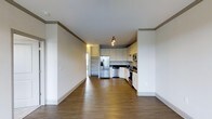 This single-floor space presents a clean, modern aesthetic defined by an open-concept layout that seamlessly connects the living area to the kitchen. The flooring throughout is a warm, medium-toned hardwood-style material that adds both warmth and cohesion to the space. Crisp white walls and bright white baseboards enhance the sense of openness and light.

The kitchen is a standout feature, outfitted with white shaker-style cabinetry extending to the upper walls, dark countertops, and a full suite of stainless steel appliances including a French door refrigerator with an ice and water dispenser, a dishwasher, a range, and an over-the-range microwave. Track lighting above the kitchen provides focused illumination, while a sink positioned along the counter offers a practical workspace. The kitchen layout is efficiently designed in an L-shape, maximizing storage and counter space.

Adjacent to the kitchen, a built-in shelving nook adds functional storage and display space. The open living area is generous and versatile, with smooth ceilings accented by crown molding that lend a refined touch to the overall design. The layout flows naturally, making the space feel bright, airy, and move-in ready.