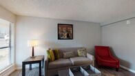 This single-floor unit offers a practical and comfortable layout designed to maximize functionality across its key living spaces. The living room serves as the central gathering area, featuring durable wood-look flooring, neutral gray walls, and a textured ceiling. It is furnished with a plush sofa accented by colorful throw pillows, a bold red accent chair, a dark wood coffee table, and a side table with a warm lamp — all complemented by a framed art piece above the sofa. A window with horizontal blinds allows natural light to fill the space.

The kitchen is outfitted with white cabinetry, a smooth black electric cooktop, an over-the-range microwave, and granite-style countertops that add a clean, polished look to the compact cooking area. A dining area is visible adjacent to the kitchen, providing a dedicated space for meals.

The bedroom is a calm retreat with the same wood-look flooring, neutral walls, a textured ceiling, and a window that brings in natural light. It comfortably accommodates a full-sized bed with coordinated bedding, a nightstand with lamp, and a dresser with ample storage. A second bedroom area is accessible through a connecting doorway.

The bathroom features a single-sink vanity with white cabinetry and a full tub/shower combination with tile surround, offering all essential amenities in a clean, well-maintained space.