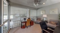 This single-floor office space offers a thoughtfully arranged and functional layout designed to accommodate both focused work and relaxed seating. The space is defined by its distinctive floor-to-ceiling glass partition walls framed in sleek aluminum, which separate distinct areas while maintaining an open, transparent feel throughout. Natural light filters in through multiple windows dressed with horizontal blinds, keeping the atmosphere bright and pleasant.

The primary work area features a circular glass-top desk paired with a woven wicker chair, complemented by a secondary workstation housing a desktop computer, monitor, and printer atop a dark wood desk with an ergonomic office chair. A bookcase with a curated selection of reading material adds a scholarly touch near the glass partitions.

A comfortable lounge area provides an inviting retreat, furnished with a cushioned armchair, a side table with a table lamp, and a recliner, making the space versatile for meetings or quiet reading. Textured popcorn ceilings with recessed lighting and ceiling fans ensure comfortable climate control and even illumination throughout. The carpet flooring in neutral tones ties all areas together cohesively. A large indoor plant adds a natural, refreshing element to the overall environment.