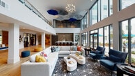 Located at Constellation Place Dr SW in Huntsville, Alabama, this single-story community clubhouse delivers a sophisticated, resort-inspired atmosphere through thoughtful architectural design and upscale interior finishes. The space is anchored by a dramatic double-height ceiling that rises above the main lounge, creating an expansive, airy ambiance accentuated by sculptural geometric ceiling fixtures in bold blue and gray tones and a striking branching chandelier. Floor-to-ceiling glass walls and black-framed French doors flood the interior with abundant natural light while offering unobstructed views of the outdoor pool courtyard, complete with teal umbrellas and landscaped seating areas. The main lounge features generous sectional and lounge seating in neutral tones accented by vibrant throw pillows, arranged atop a bold starburst-patterned area rug over warm-toned hardwood flooring. A textured wood-paneled accent wall frames a bar-style island with a marble countertop and gold-accented stools, serving as a functional social hub. A separate media lounge area with plush blue velvet chairs and a wall-mounted television provides additional gathering space. A glass-enclosed fitness center with blue framed doors houses cardio and strength equipment with natural light from exterior windows. An upper mezzanine level, visible behind glass railings, overlooks the main floor, adding architectural depth to the overall design.