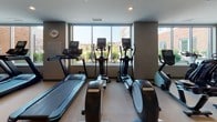This fitness center offers a thoughtfully designed, single-level workout facility that caters to a wide range of exercise needs. The space is divided into distinct functional zones, providing an organized and efficient layout for users.

The cardio area is positioned along a wall of large windows, allowing natural light to flood the space and offering pleasant outdoor views of the surrounding landscape. This section is equipped with multiple treadmills, elliptical trainers, and a stair climber, all arranged to take advantage of the natural light and views.

The strength training area features a variety of cable machines and weight equipment, including multi-functional cable towers and seated weight machines, providing options for a full-body workout. Floor-to-ceiling mirrors line one wall, enhancing the sense of space and allowing users to monitor their form during exercises.

The facility is finished with durable, light-toned rubber flooring throughout, ensuring both safety and comfort. Recessed lighting in the smooth, neutral-toned ceiling provides consistent illumination across the entire space. A wall-mounted flat-screen television adds an entertainment element to the environment. The overall ambiance is clean, modern, and functional, making it a practical and inviting space for fitness activities.