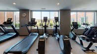This fitness center offers a thoughtfully designed, single-level workout facility that caters to a wide range of exercise needs. The space is divided into distinct functional zones, providing an organized and efficient layout for users.

The cardio area is positioned along a wall of large windows, allowing natural light to flood the space and offering pleasant outdoor views of the surrounding landscape. This section is equipped with multiple treadmills, elliptical trainers, and a stair climber, all arranged to take advantage of the natural light and views.

The strength training area features a variety of cable machines and weight equipment, including multi-functional cable towers and seated weight machines, providing options for a full-body workout. Floor-to-ceiling mirrors line one wall, enhancing the sense of space and allowing users to monitor their form during exercises.

The facility is finished with durable, light-toned rubber flooring throughout, ensuring both safety and comfort. Recessed lighting in the smooth, neutral-toned ceiling provides consistent illumination across the entire space. A wall-mounted flat-screen television adds an entertainment element to the environment. The overall ambiance is clean, modern, and functional, making it a practical and inviting space for fitness activities.
